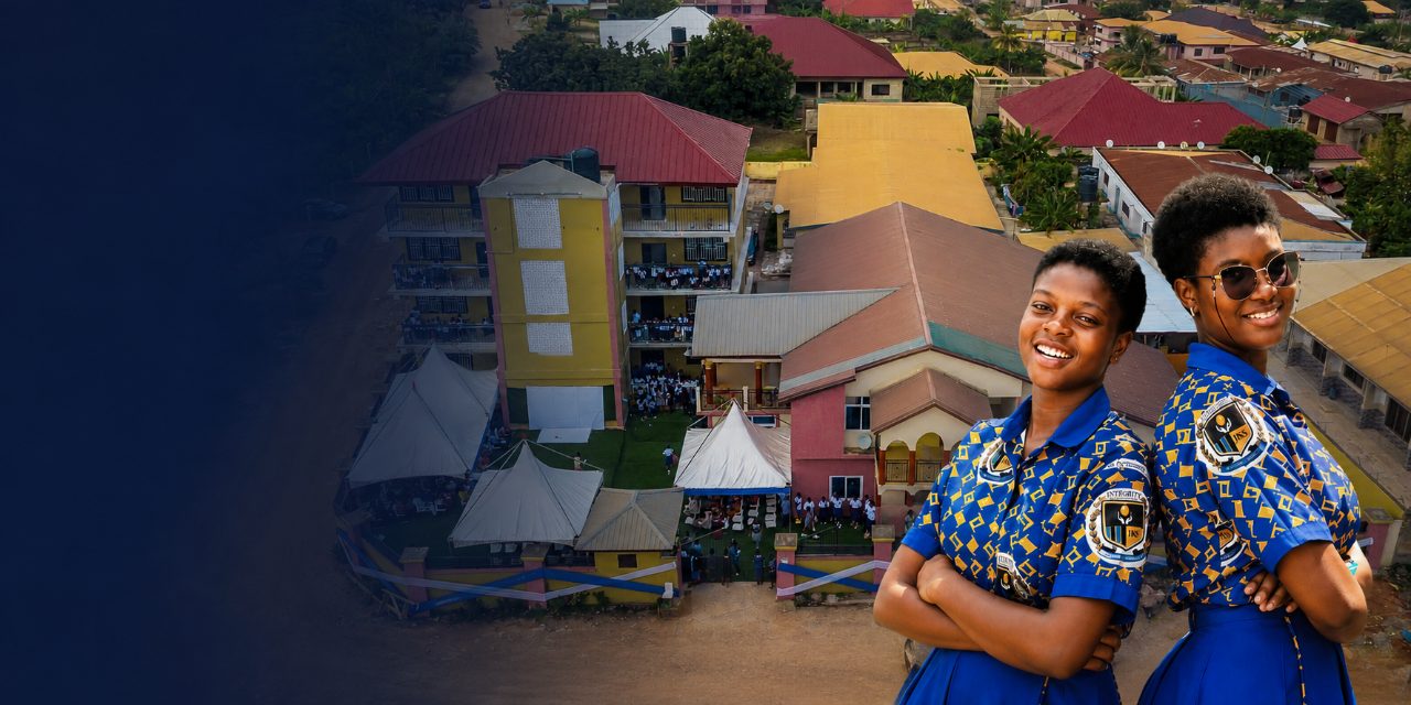 Two Integrity Keepers' School students in school uniform, with an aerial view of the school campus and the surrounding Asuoyeboah North neighbourhood in Kumasi behind them.
